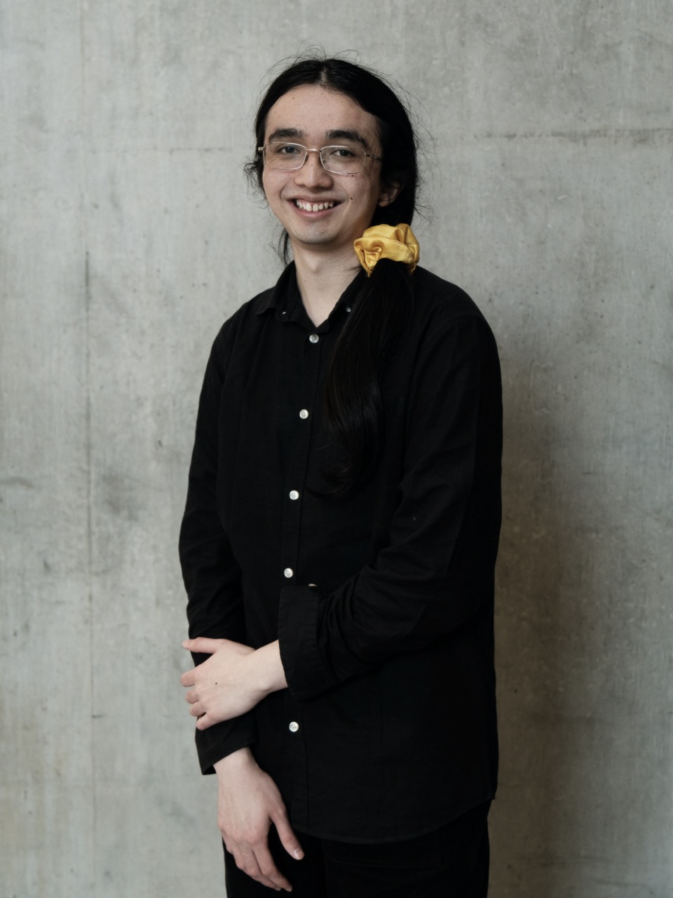 A photo of Jack wearing a black shirt and a yellow scrunchie, standing in front of a concrete wall